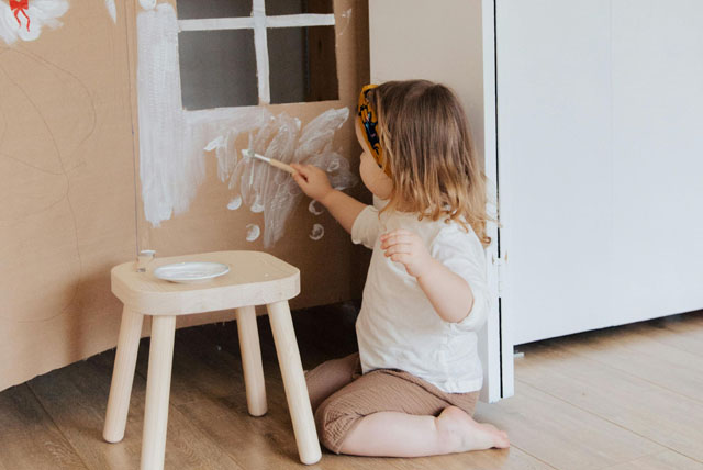 toddler girl painting white paint cardboard house